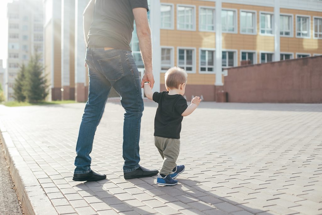 Un papà cammina in città tenendo per mano il suo bambino, ripresi di spalle.