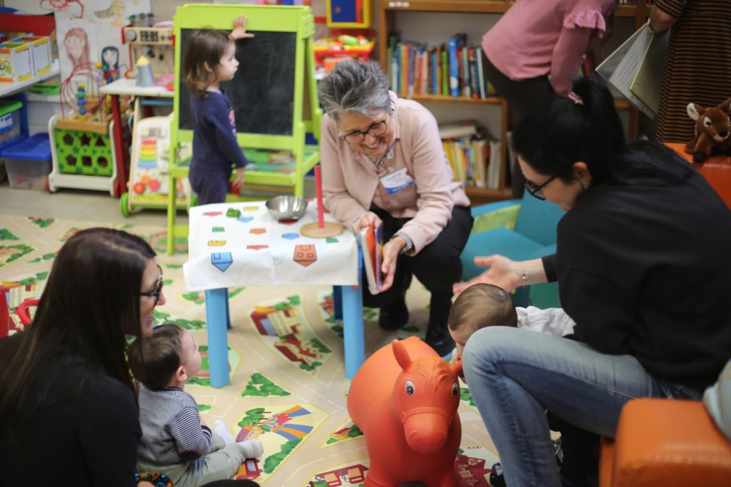 Una volontaria legge un albo a bambine e bambini piccoli insieme alle persone adulte che li accompagnano.