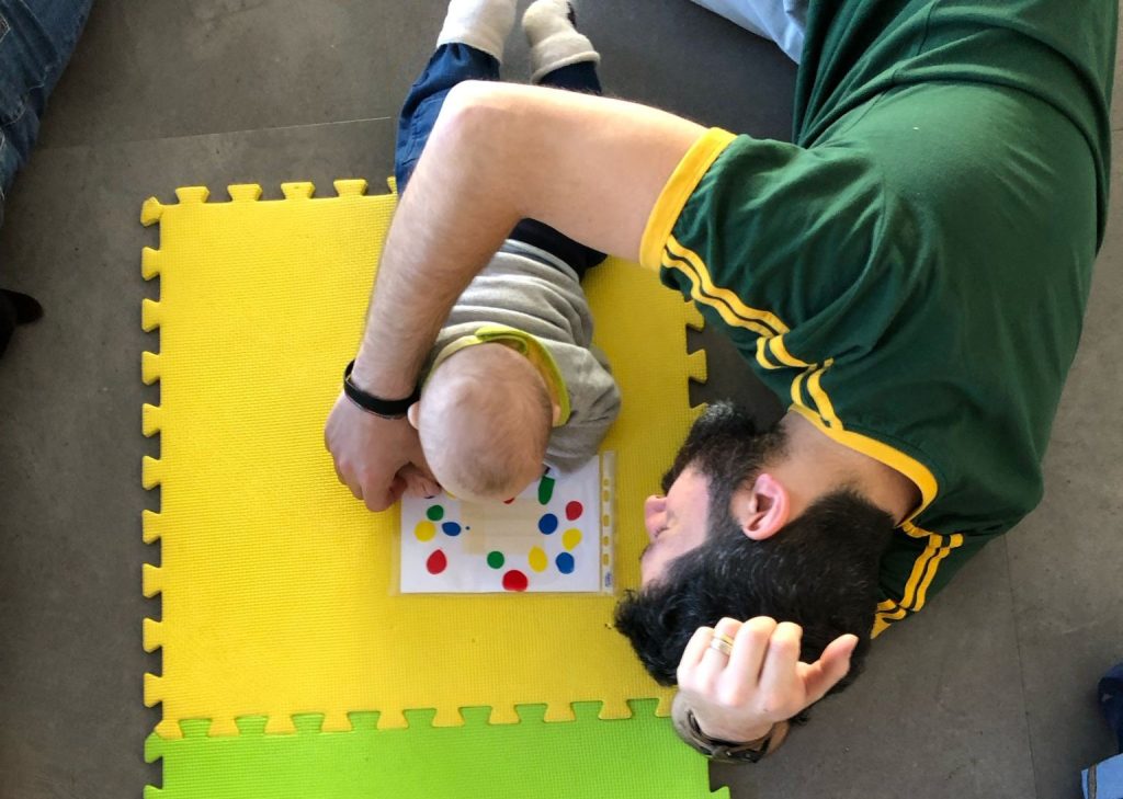 Un adulto sdraiato accanto a un bambino su un tappeto giallo osserva con lui una scheda colorata.