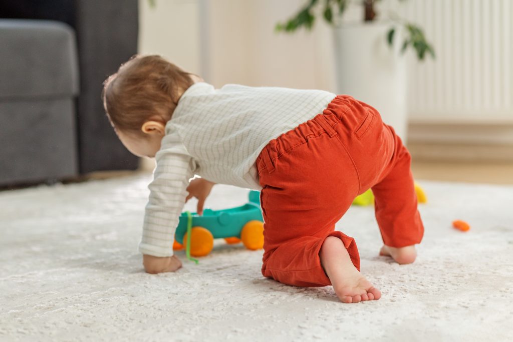 Un bambino con i pantaloni rossi si muove a quattro zampe sul tappeto, vicino ai suoi giochi.