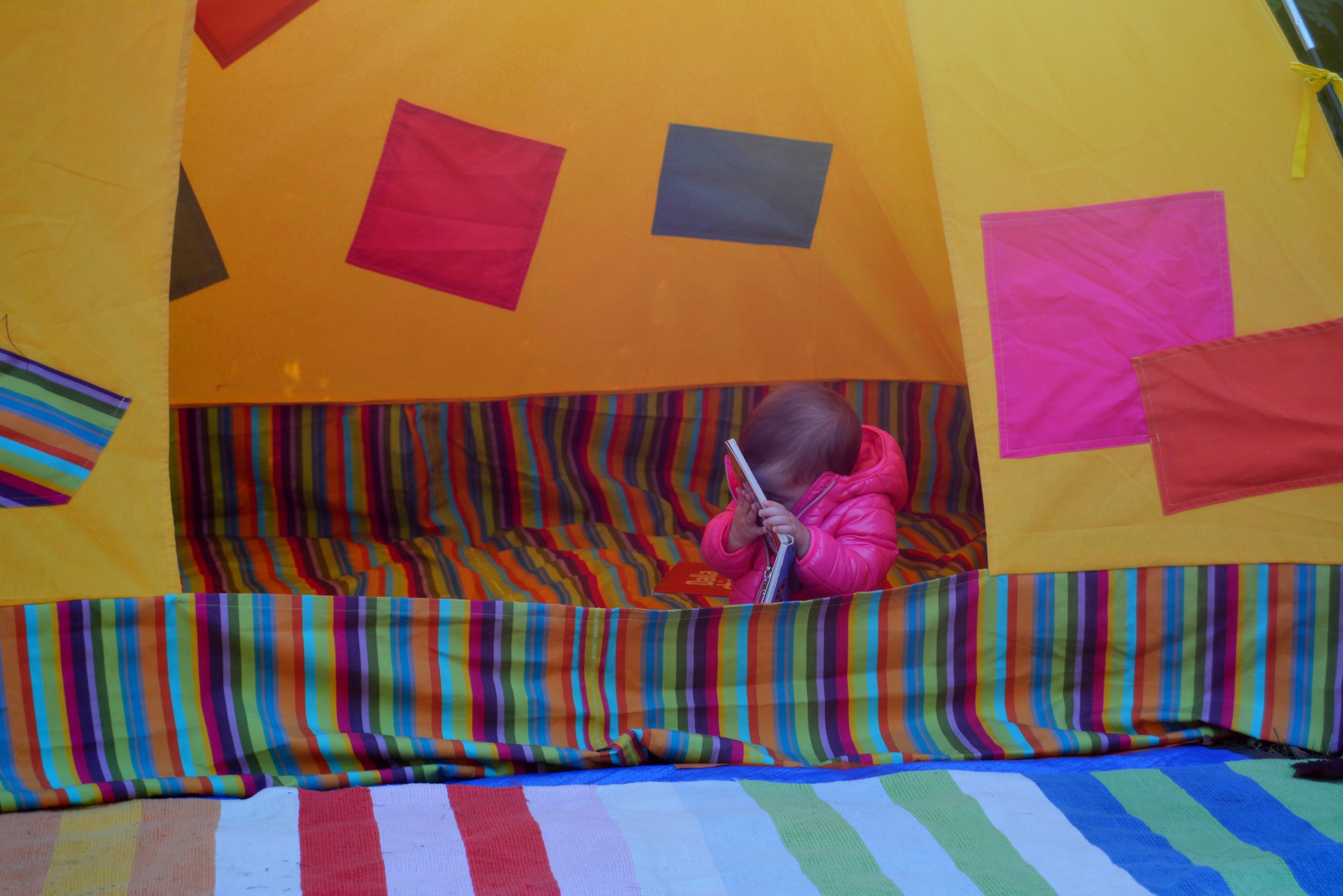 Una bambina piccola dentro una tenda colorata tiene un libro cartonato davanti al viso