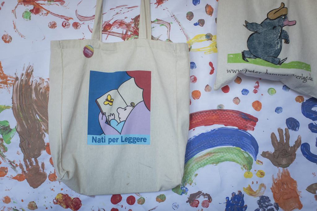 Una borsa di tela di Nati per Leggere appoggiata su un telo dipinto con impronte di mani colorate