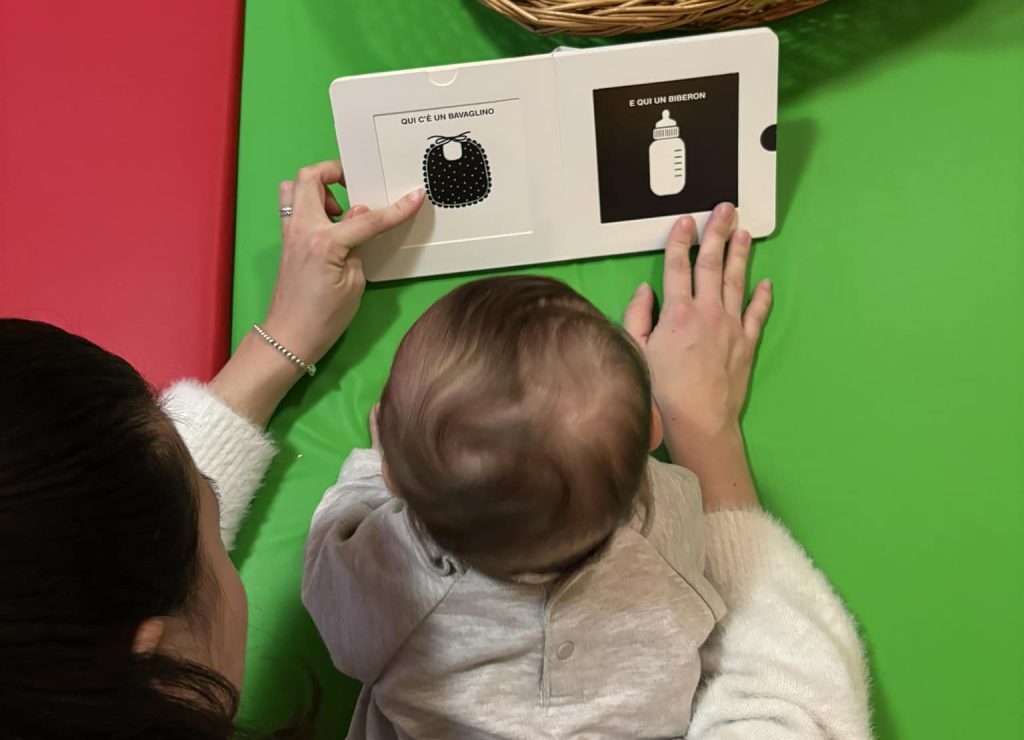 Una neonata o un neonato a pancia in giù guarda un libro cartonato in bianco e nero ad alto contrasto, accanto a un cesto di primi libri