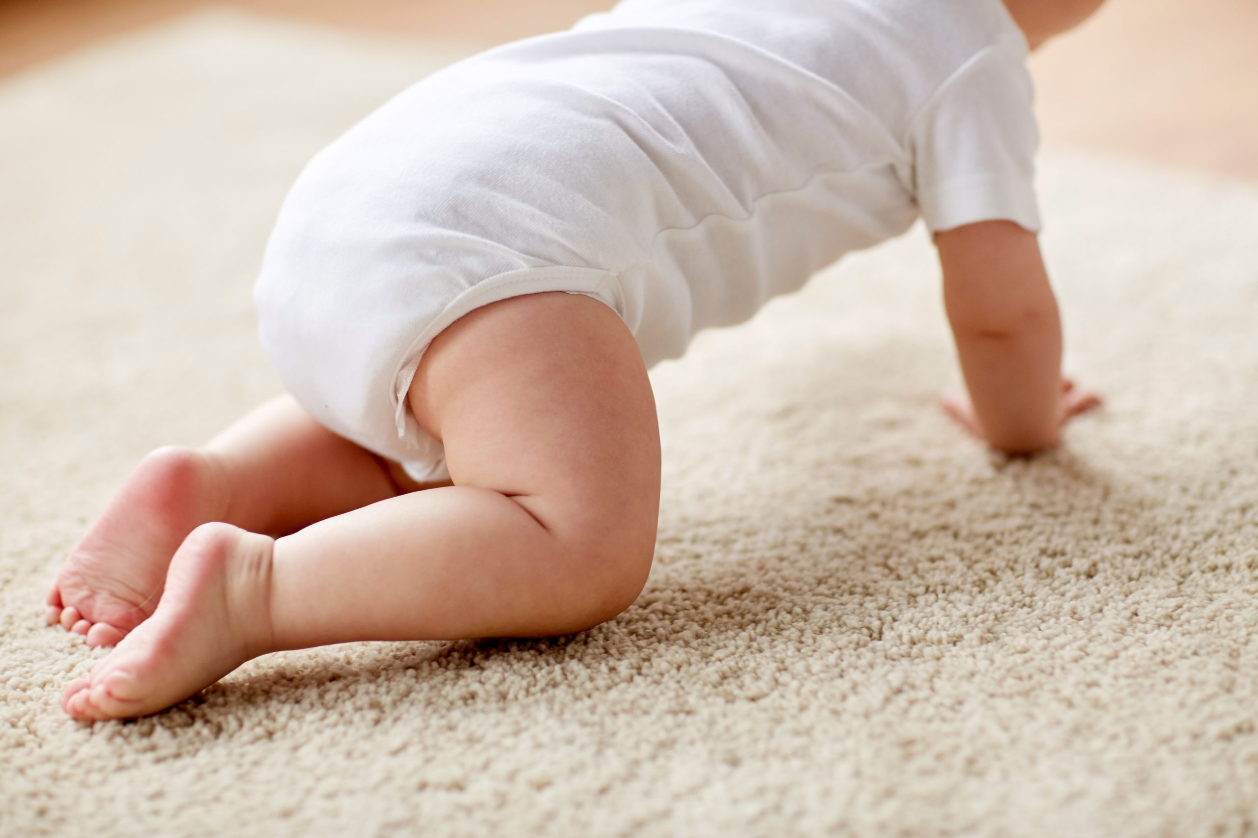 Un bambino in tutina bianca gattona a quattro zampe su un tappeto chiaro.